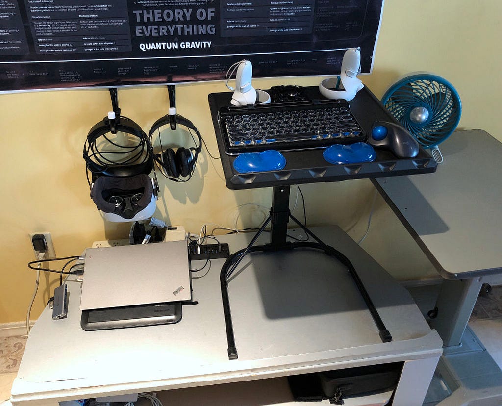A raised keyboard tray on a low stand, which also holds a closed laptop computer. A VR headset and pair of headphones hang from hooks on the wall next to the keyboard.