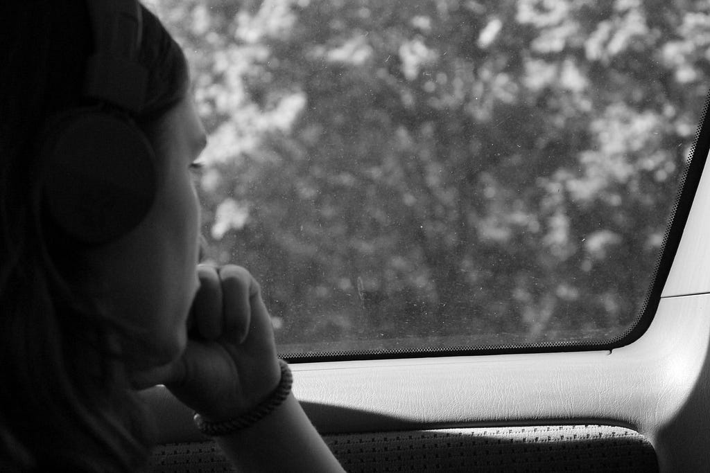 ID: Black and white photo of a long-haired person in profile. They’re looking out of a car window and wearing headphones.