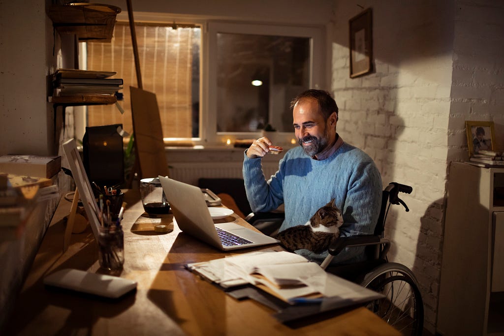 A man in a wheelchair sits at a desk in a home office smiling at his computer with a cat on his lap.