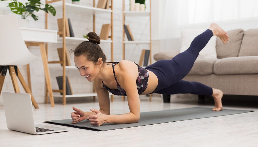 Woman doing a yoga pose, dressed in exercise wear. She is looking at a laptop in front of her. She is smiling.
