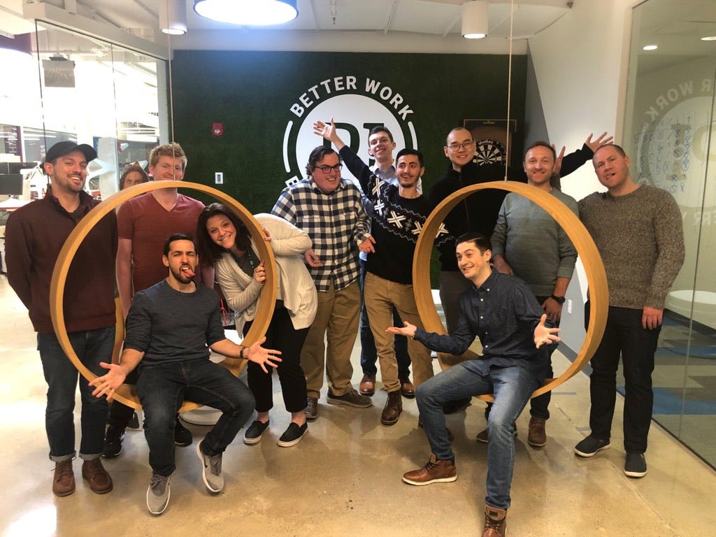 Two men sit in circular swings suspended from the ceiling while a group of people surround them, smiling at the camera.