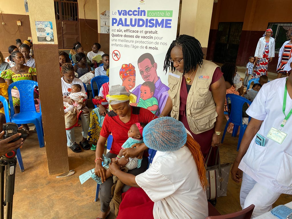 A health care worker in a scrub cap prepares to administer a vaccine to a young girl sitting in an adult woman’s lap as a USAID staffer looks on. They are in front of a sign that says “Paludisme,” which is French for malaria.