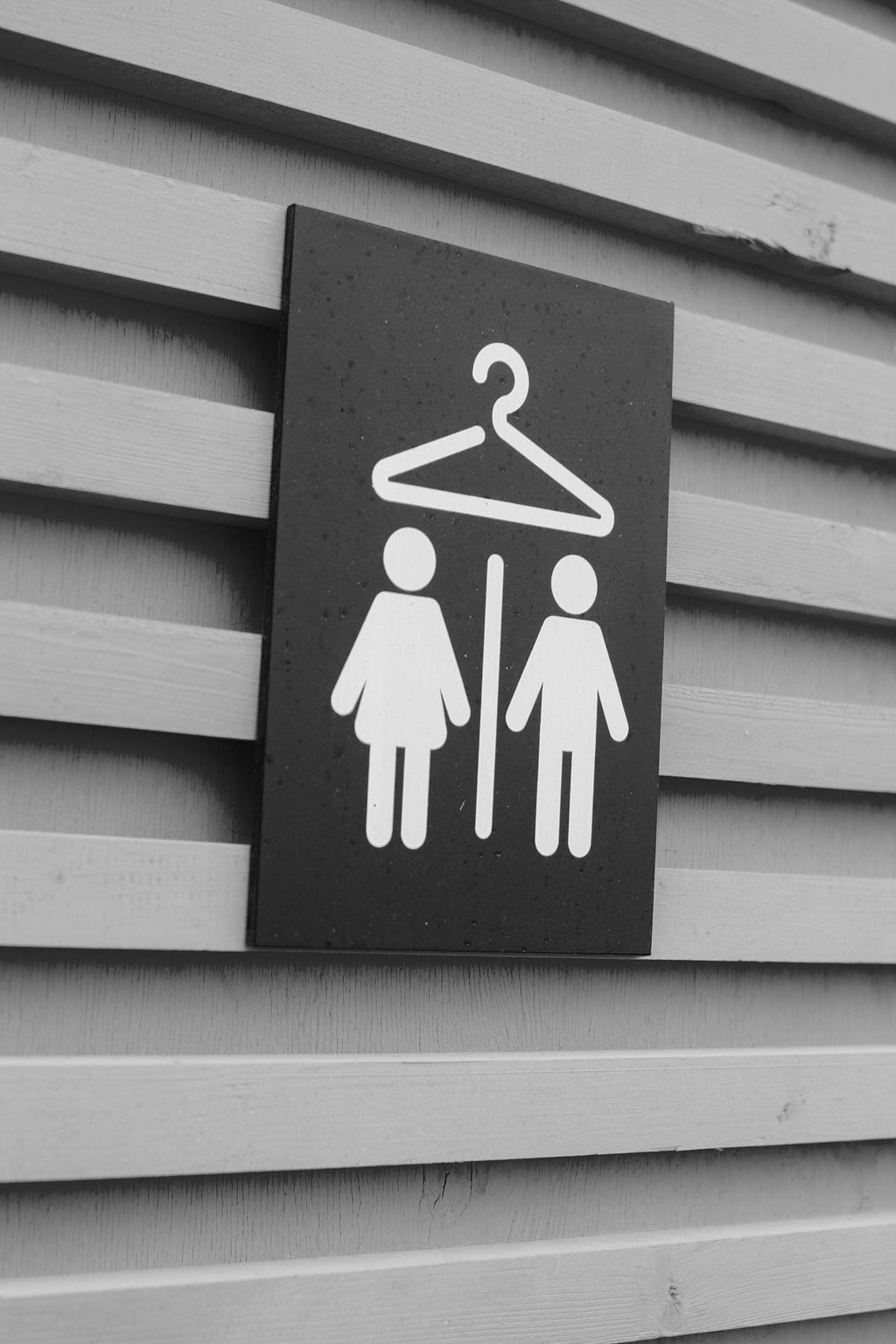 A displaying a pair of stick-figure gender symbols — female on the left and male on the right — separated by a line and a clothes hanger above, indicating a sex-segregated pair of changing rooms.
