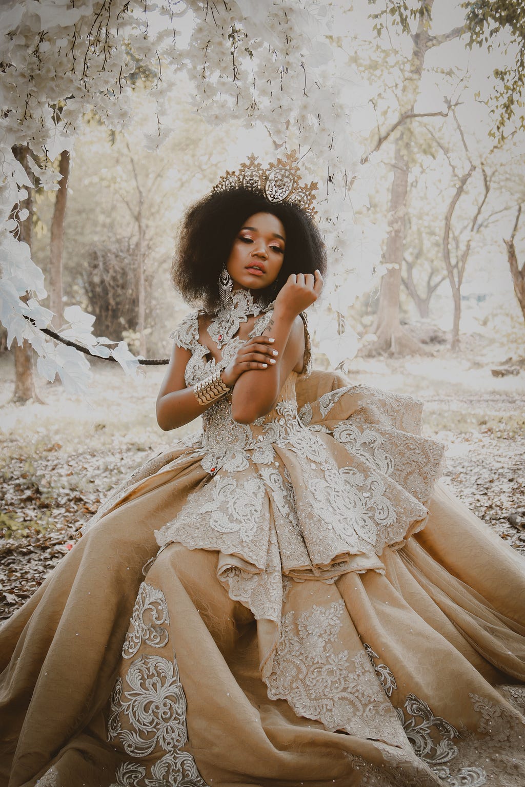 Woman with brown skin and long, textured hair wearing a taupe-toned wedding dress with elaborate white lace embellishments, against an icy winter forest background.