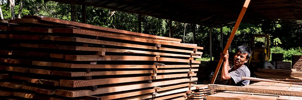 A worker lifts a piece of wood onto a  tall stack of lumber sitting underneath a metal-roofed shelter in an FSC-certified forest in Guatemala.