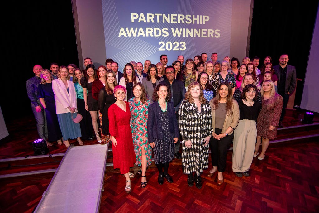 A group photograph of all the Partnership Awards winners stood on the wooden stage in the Leeds University Union Refectory at the end of the celebration.