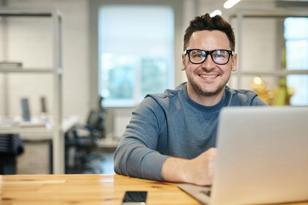 A white man with black glasses sitting at a laptop in a modern office with a dopey smile on his face. He’s confused why you sound like a click-clack typewriter.