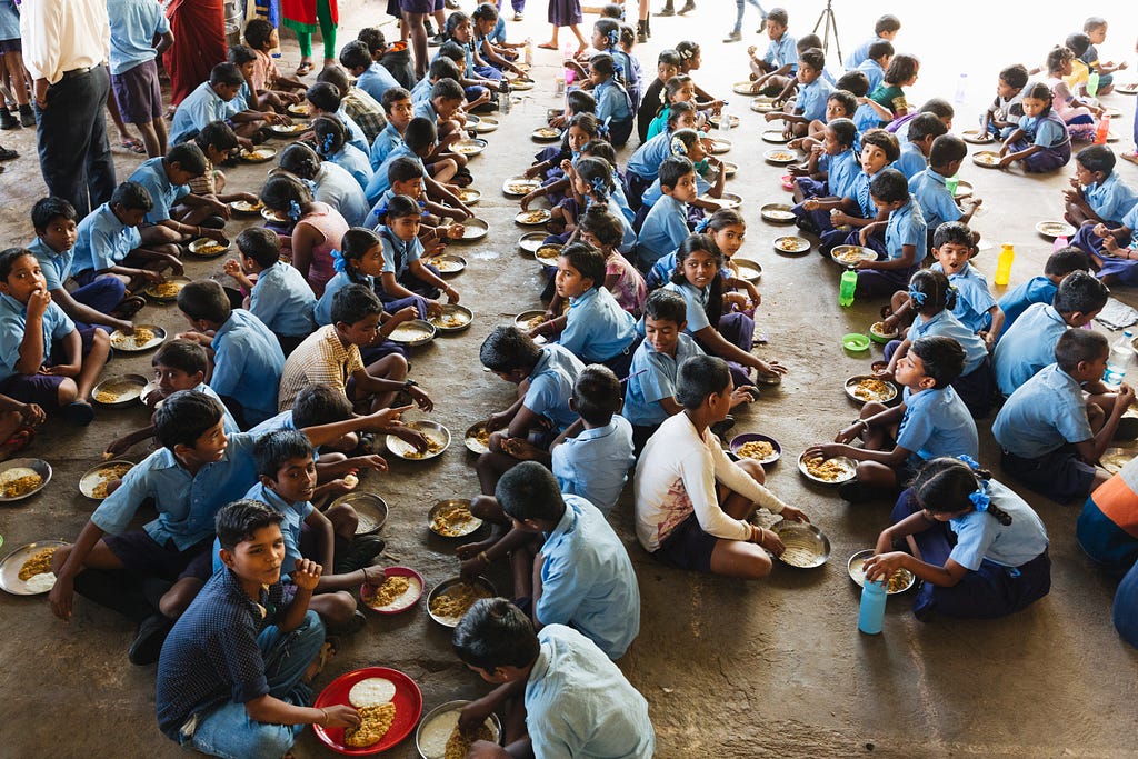 A large group of school children in matching uniforms and lined up on a floor in seated positions with plates of food in front of each one of them.