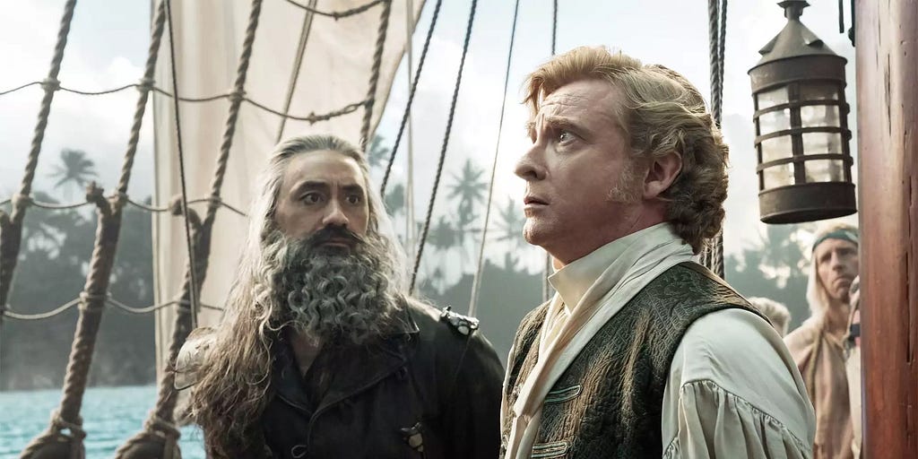 Taika Waititi as Blackbeard looks at Rhys Darby as Stede Bonnet, while Stede looks away with a fearful expression. Blackbeard is in all leather and Stede is wearing an 18th-century outfit with a white blouse and vest. They are on a ship.