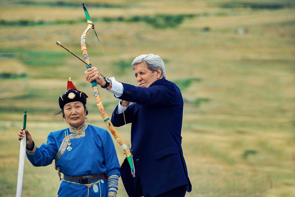 U.S. Department of State: “Archers in traditional outfits guide U.S. Secretary of State John Kerry as he prepares to shoot an arrow while attending a “mini-nadaam” — a display of music, dance, archery, wrestling, and horse-riding — in a field outside Ulaanbaatar, Mongolia, following bilateral meetings on June 5, 2016, with Mongolian President Tsakhia Elbegdorj and Foreign Minister Lundeg Purevsuren.”