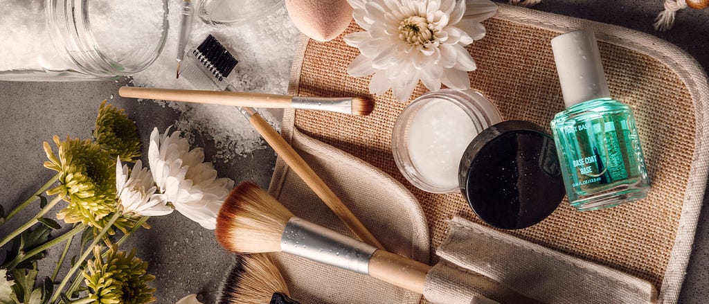 Big and small makeup brushes, spilled bath salts, green and white chrysanthemums, a bottle of transparent blue nail polish, and other beauty products lay flat on a gray table-top and an open beige makeup case.
