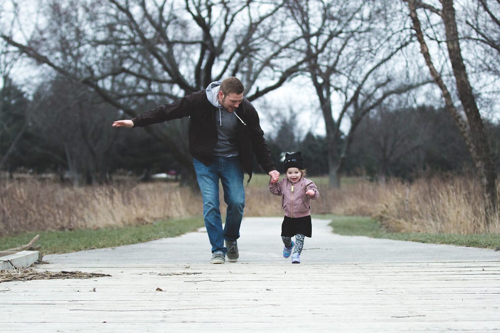 Pai corre de mãos dadas com a filha bebê em um bosque repleto de árvores. A criança sorri, feliz. Esta foto ilustra um texto sobre como obter residência permanente na Estônia.