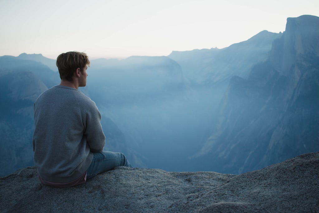 a man sitting on the edge of a rock, looking over a massive and foggy valley