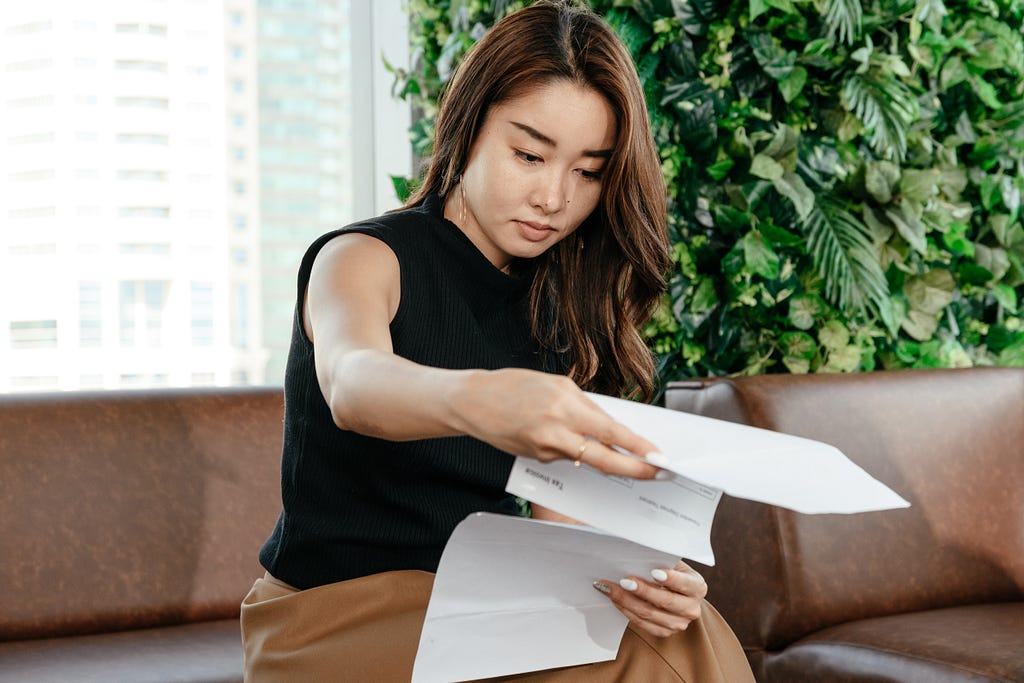 A woman wearing a black blouse and brown skirt is sitting on a brown leather couch in front of a window and a large plant with green foliage. She is looking through paperwork.