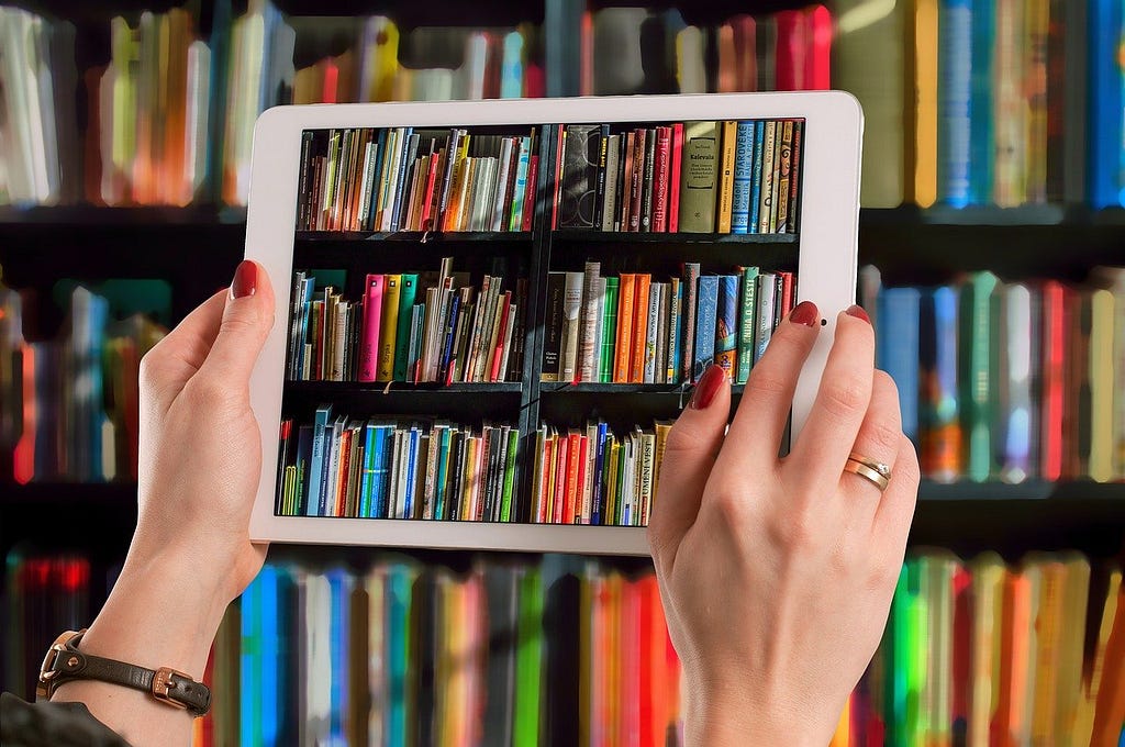 A person is holding up a tablet in front of a bookcase filled with books. They are taking a picture with the tablet, so the books are visible on the tablet screen as well. Only the person’s hands are visible. They wear read fingernail polish and have a ring on their right ring finger. They also wear a watch on their left wrist.