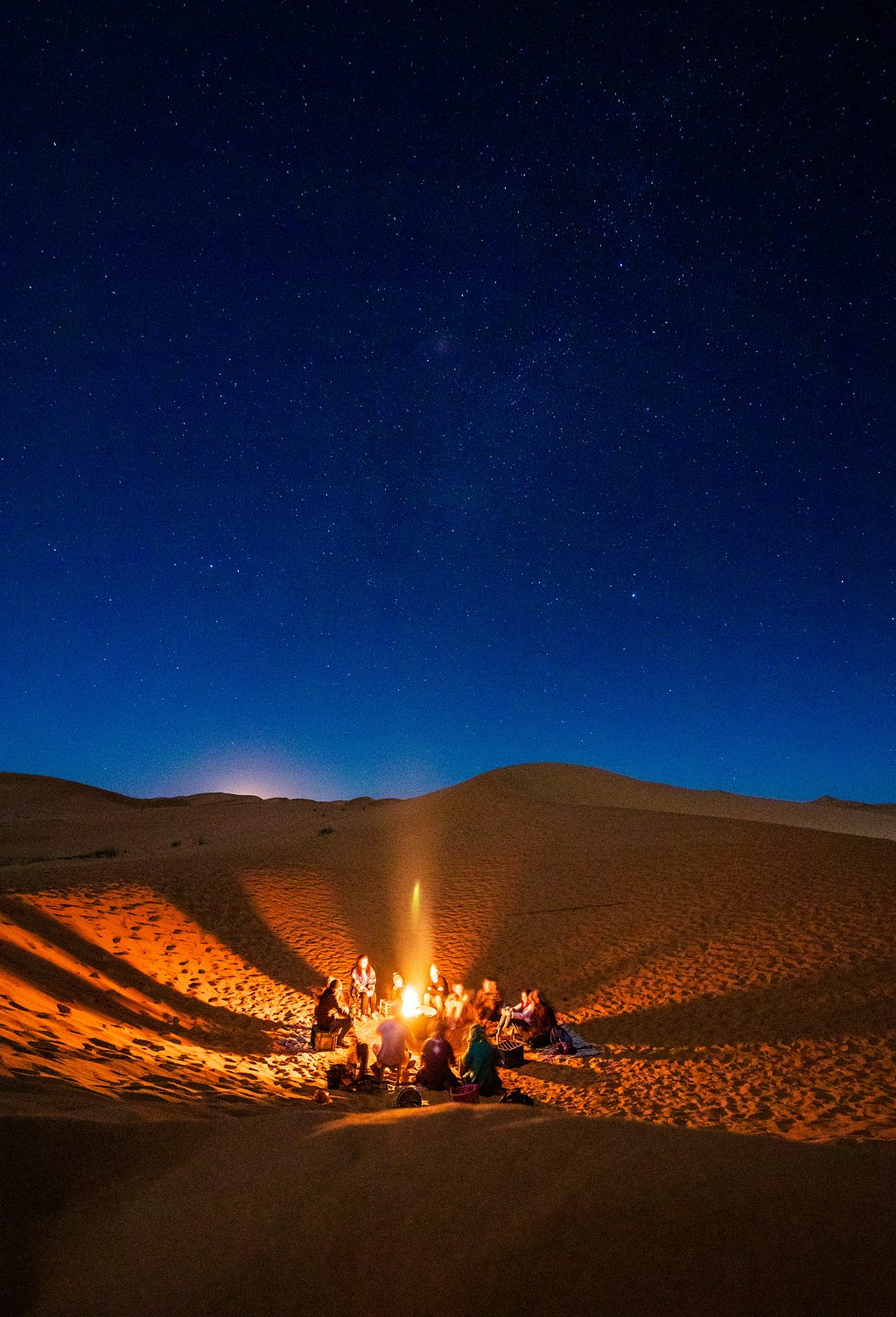 A night sky in the desert with people sitting around a campfire.