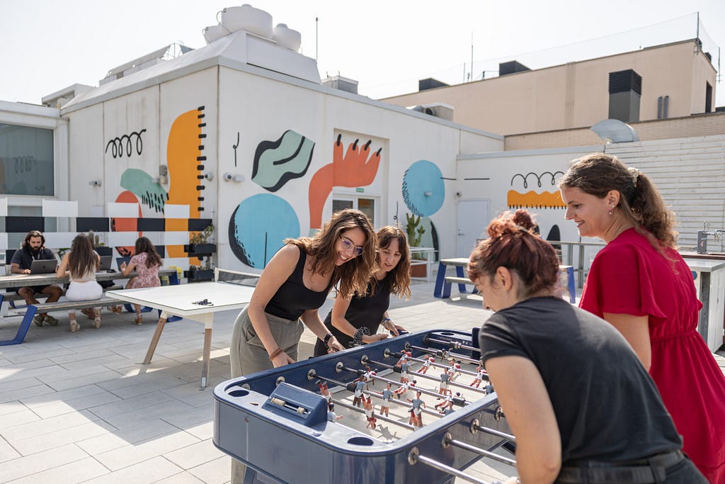 Photograph: In the foreground female colleagues play table football on the sunny rooftop terrace of Qonto’s offices in Barcelona. In the background other colleagues are seen seated at picnic tables. The walls have brightly colored mural paintings.