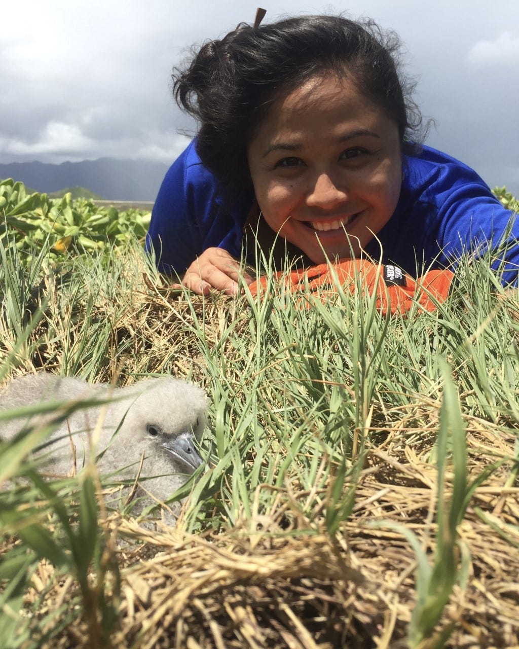 Smiling person in blue shirt lays on grass next to seabird chick.