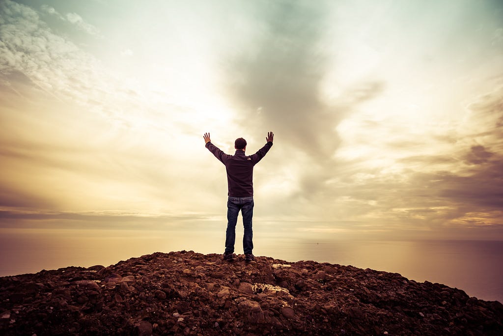 This is a man who is standing on a rock. The colour of the rock is dark brown. The man is standing in front of the sea. We can see clouds in the background. The man is raising his hands in front of the sky. It seems he is admiring the beauty of nature.