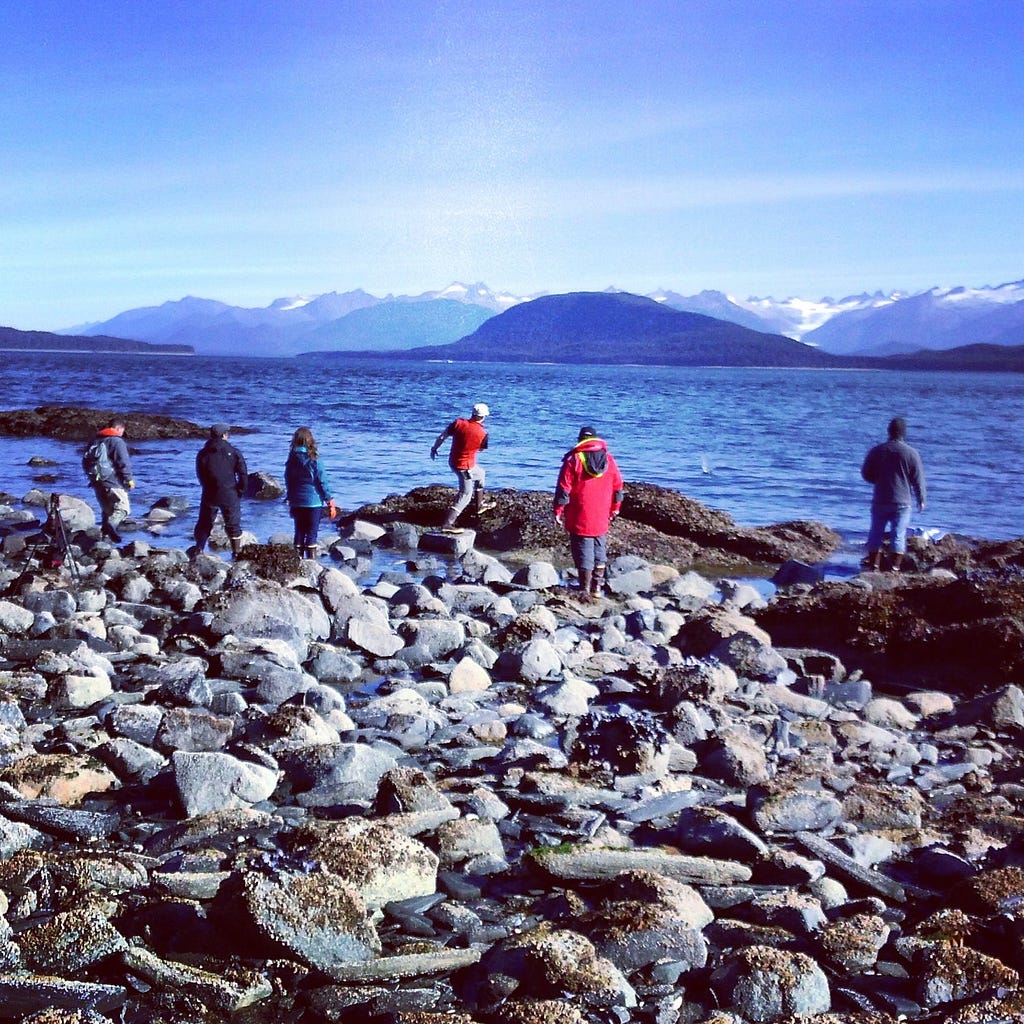 photo of six people with their back towards the camera, all adults appearing to be having fun skipping rocks on beautiful navy blue water where snow capped mountains can be seen in the background. two of the people have on bright red jackets which makes all the colors seem more harmonious and beautiful together. they are all dressed in winter gear.