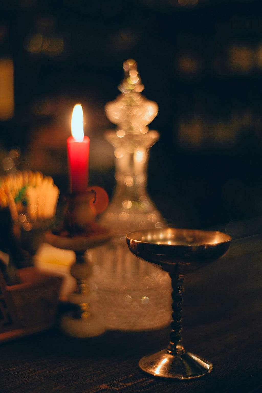 Photo of a golden chalice on a dark surface. Behind it, the image blurs, showing a lit, red tapered candle in candleholder, and an ornate crystal carafe with a stopper. A few other items are blurred and in the background, without being clearly identified. The darkness of the photo feels intimate, sacred, or of private importance.