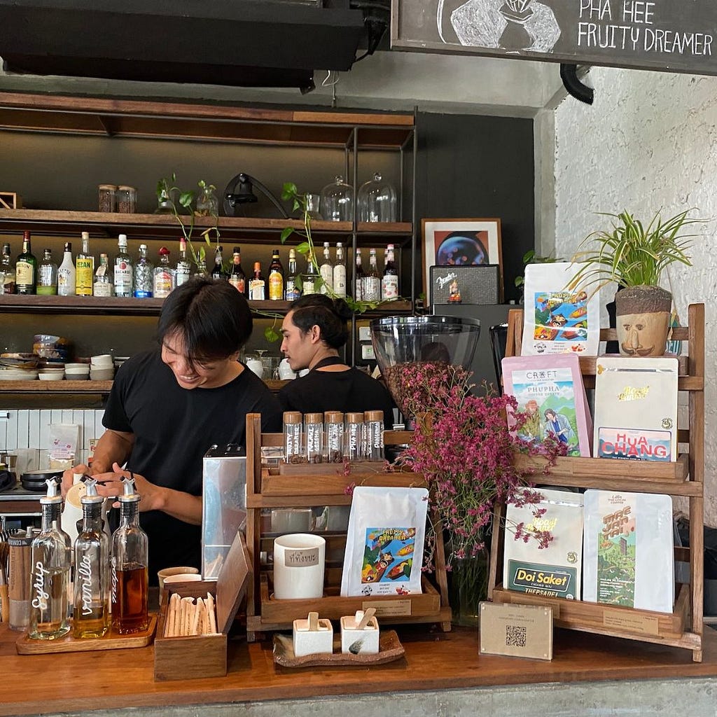 Two young baristas, one smiling,  at work behind a cafe bar. Bottles of alcohol line the shelves in background, colourful bags of coffee in foreground.