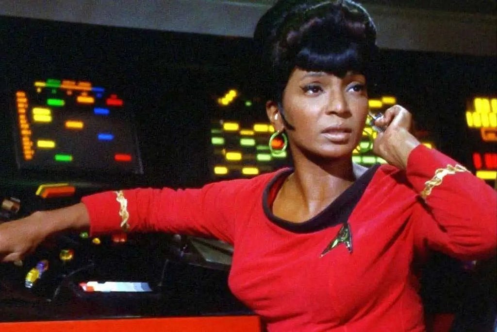 Nichelle Nicolls looking absolutely radiant as Lieutenant Uhura in Star Trek: The Original Series. She is wearing a red uniform, with her hair in an immaculate beehive, as she sits by her comms station. She has one hand at her earpiece as she gaze off to the right, with a look of deep concentration on her face.