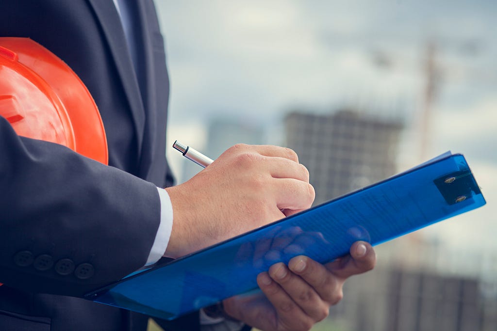 Person in a suit with a hard hat writing on a blue clipboard