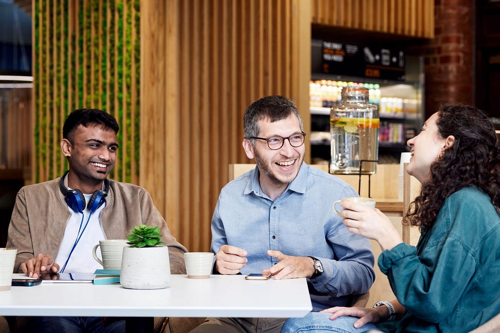 Three students sit enjoying a drink in a cafe on campus. They are smiling and laughing. One is holding a cream cup, one is sat with a tablet on the table in front of them and the third with a mobile phone on the table in front of them. There is a plant in white holder on the table.