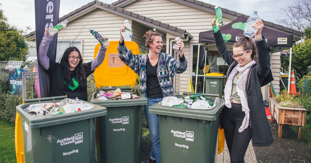 Three women standing behind three council recycle bins laughing and holding up plastic bottles