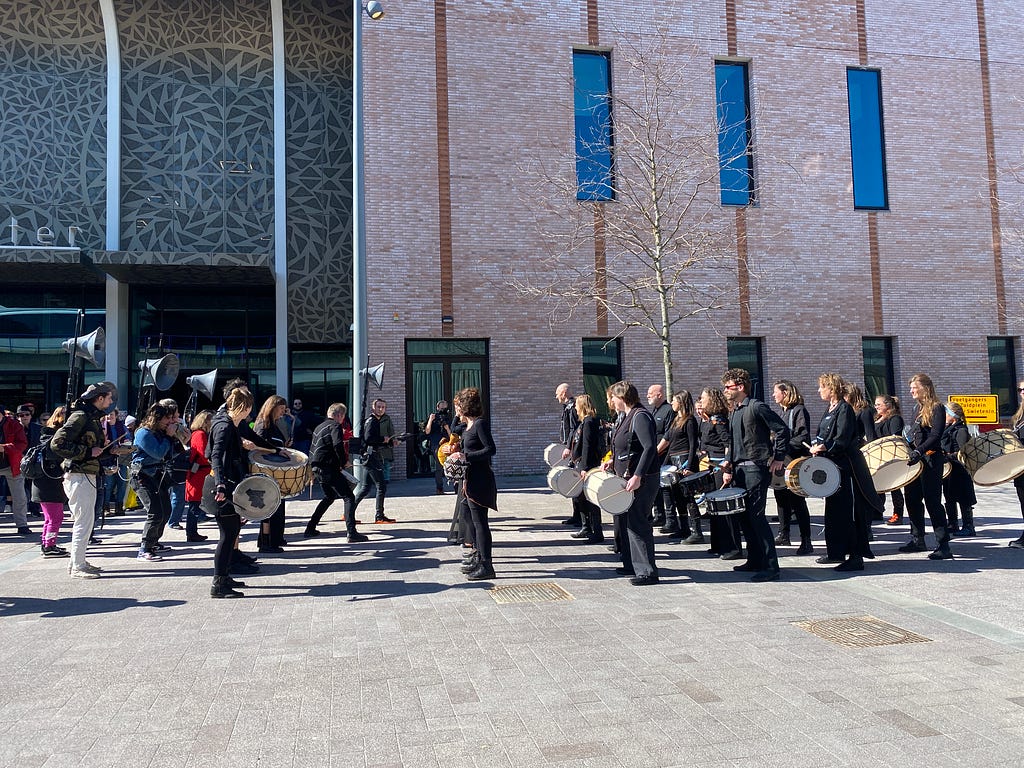 Two groups of percussionists face each other, one group carrying tall speakers on their backs, outdoors in front of a Theatre.
