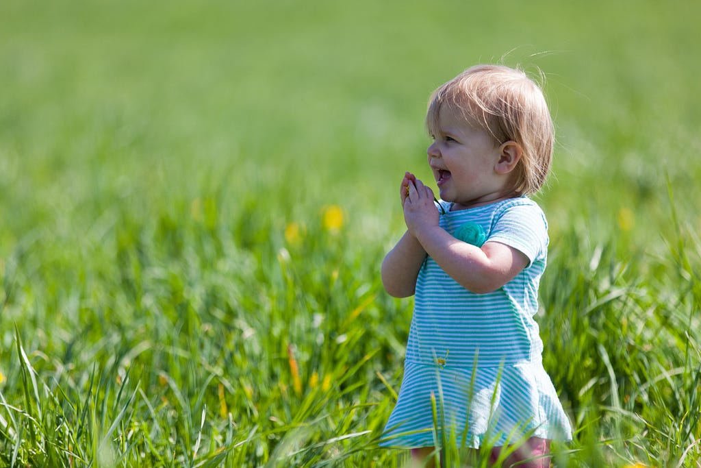 A little happy girl clapping while standing in field with grass around her by Abhay Gautam