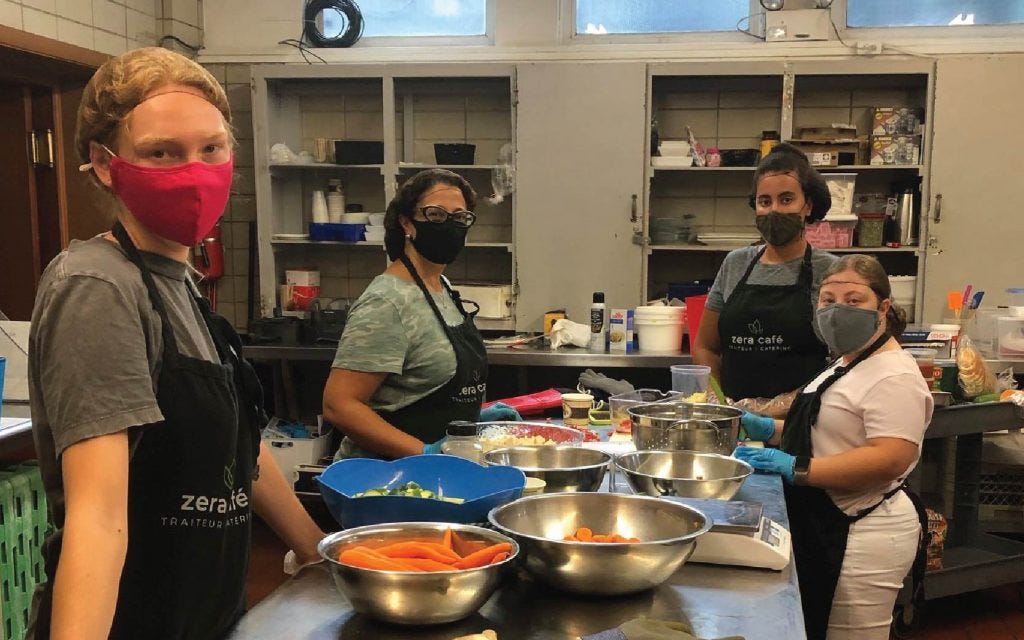 Four women in masks and apron are prepping food in the catering kitchen.