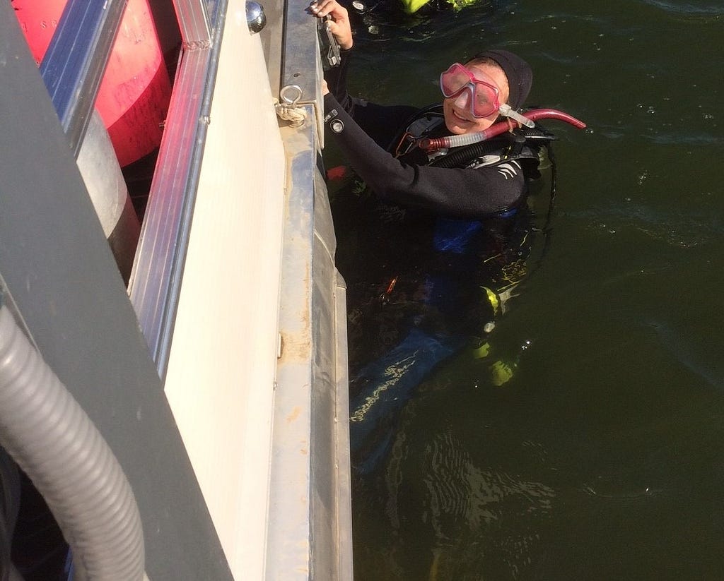 A women in scuba equipment is halfway submerged in water and holds onto the edge of a boat.