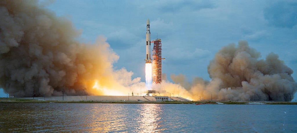 Photograph of Saturn V seconds after launch in 1973. The rocket and tower are across a watery expanse, and clouds of fire and smoke burst all around it.
