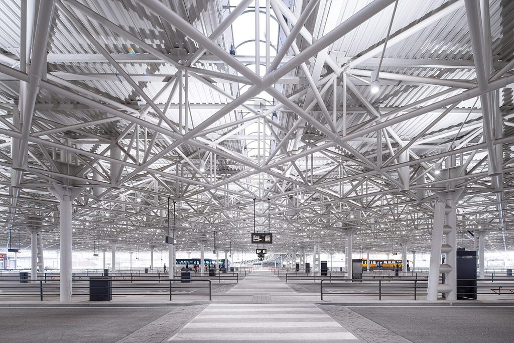 The interior of the Zvonarka Bus Terminal. The white, crisscrossed steel structure of the roof takes up the majority of the image due to its dramatic low angle. The grey, concrete floor also contains a white crossing moving away from the camera. Signs and bins are spread across the floor.
