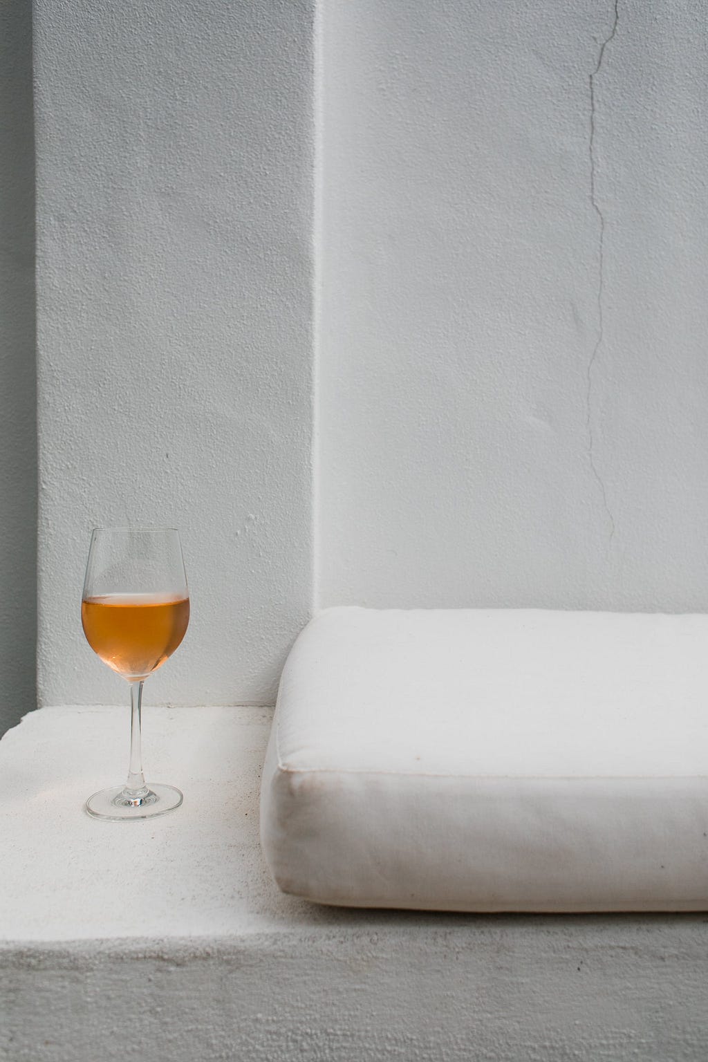 A glass of white wine next to a white wall and white pillow.