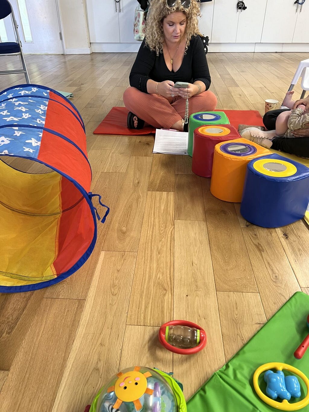 A woman with curly blonde hair sits cross-legged on a wooden floor, looking at her phone. Colorful children’s toys are scattered around, including a play tunnel, stacking cups, and soft blocks. The setting appears to be an indoor play area or childcare facility.