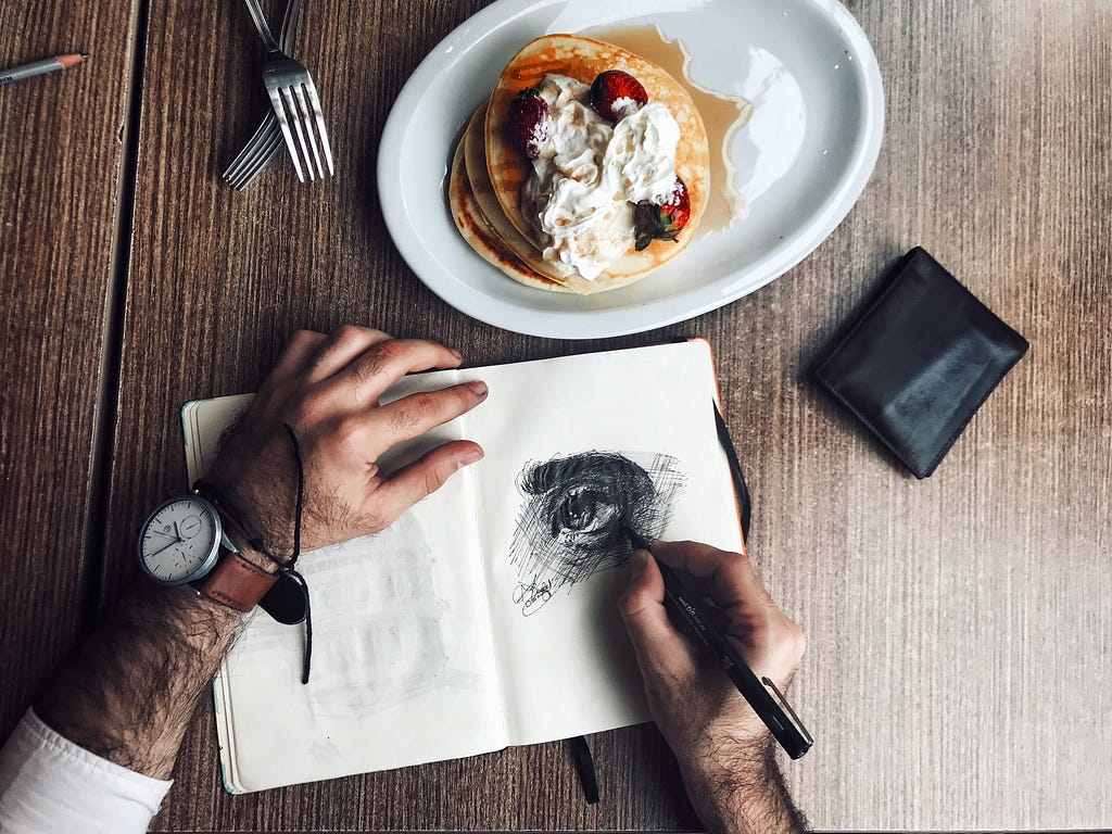 Man drawing an eye in the notebook sitting at the table with food