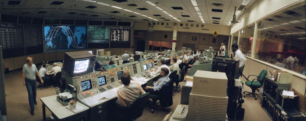 Wide angle view of the flight control room (FCR) of the Mission Control Center (MCC). Some of the STS 41-G crew can be seen on a large screen at the front of the MCC along with a map tracking the progress of the orbiter. It’s pretty intense!