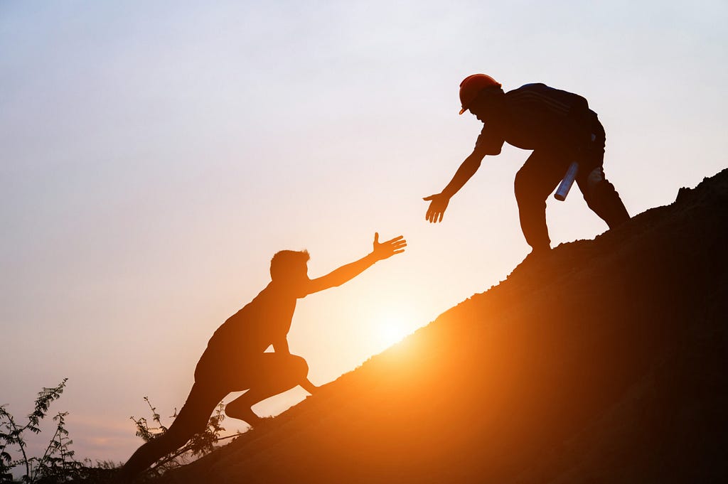 Tourists go up the hill in the sunrise, with one extending a hand to the other who is slightly below him.