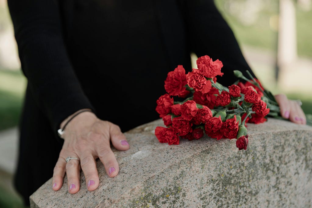 A photo shows the top of a gravestone. Behind the gravestone the torso of a person is shown in black clothing. The person’s hands are resting on the gravestone. Their left hand holds a bouquet of red flowers that are also resting on the gravestone. The person wears pink polish on their fingernails and they wear a ring on their right hand and a bracelet on their right wrist.