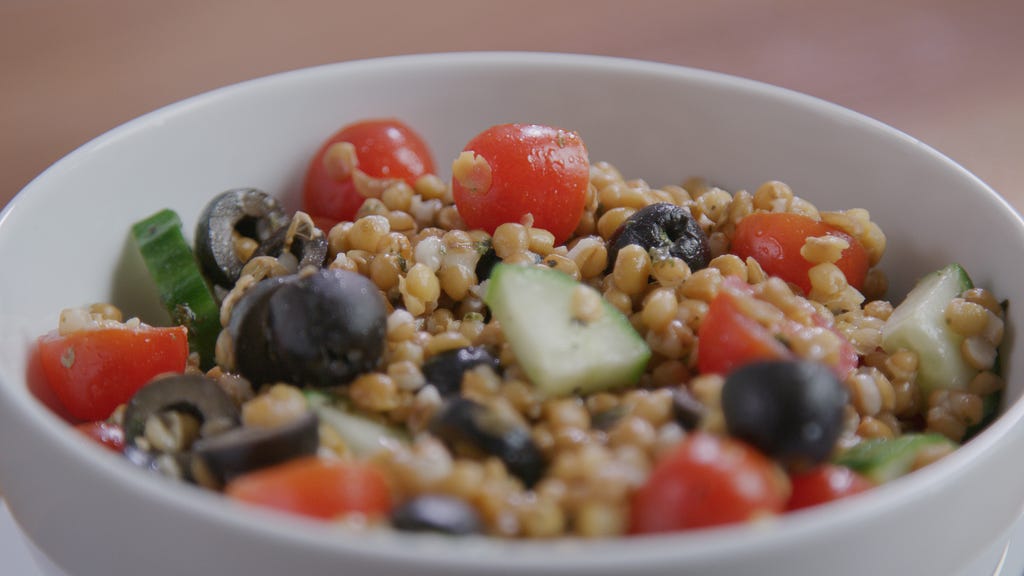 A close up of a wheat berry and lentil salad in a white bowl with cut cherry tomatoes, cucumbers and black olvies.