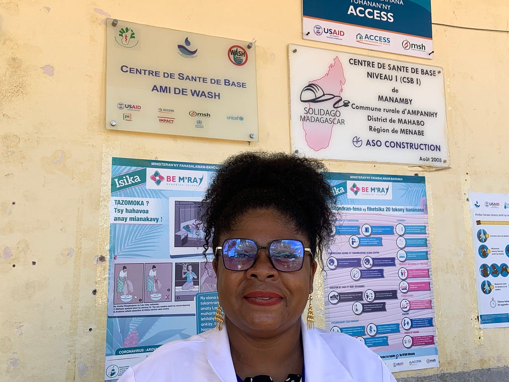 A nurse-midwife wearing stylish glasses and a lab coat poses next to a wall of health reminders posted at the health care facility where she works.