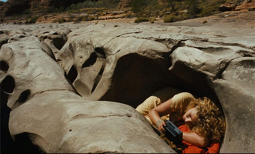 Still image showing a woman in a yellow dress lying hidden among large rocks, staring at a black electonic device