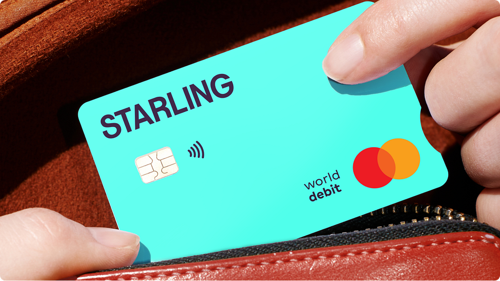 A bright teal Starling debit card with the word “STARLING” in bold purple-black, being slid into a brown leather wallet.