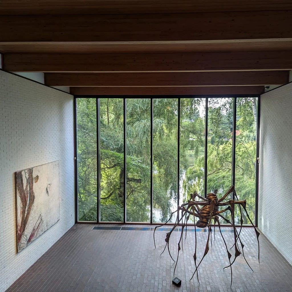 A view of a gallery room with a floor to ceiling window that looks out at trees and a lake. On the left is a large abstract painting in black and white, and to the right is a large metal sculpture of a spider.