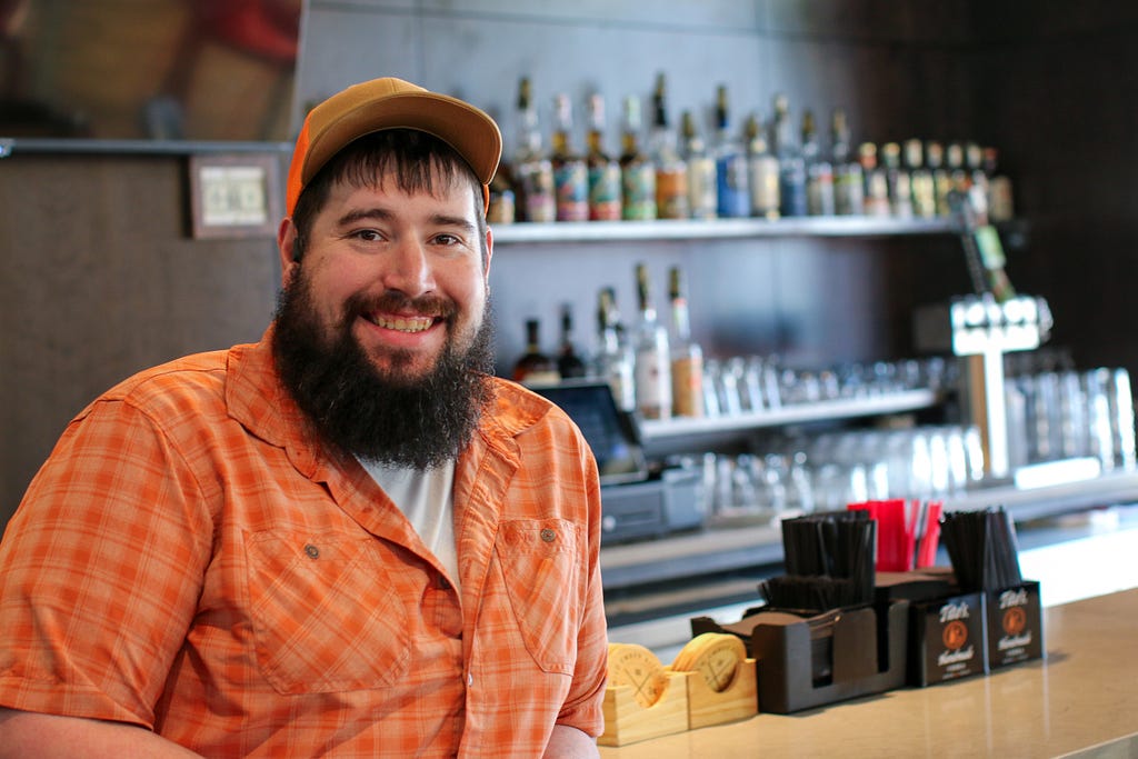 A man leans against a bar top with liquor bottles, beer tap and glassware visible behind him. He’s wearing a green shirt and ball cap, has a full beard, and is smiling at the camera.