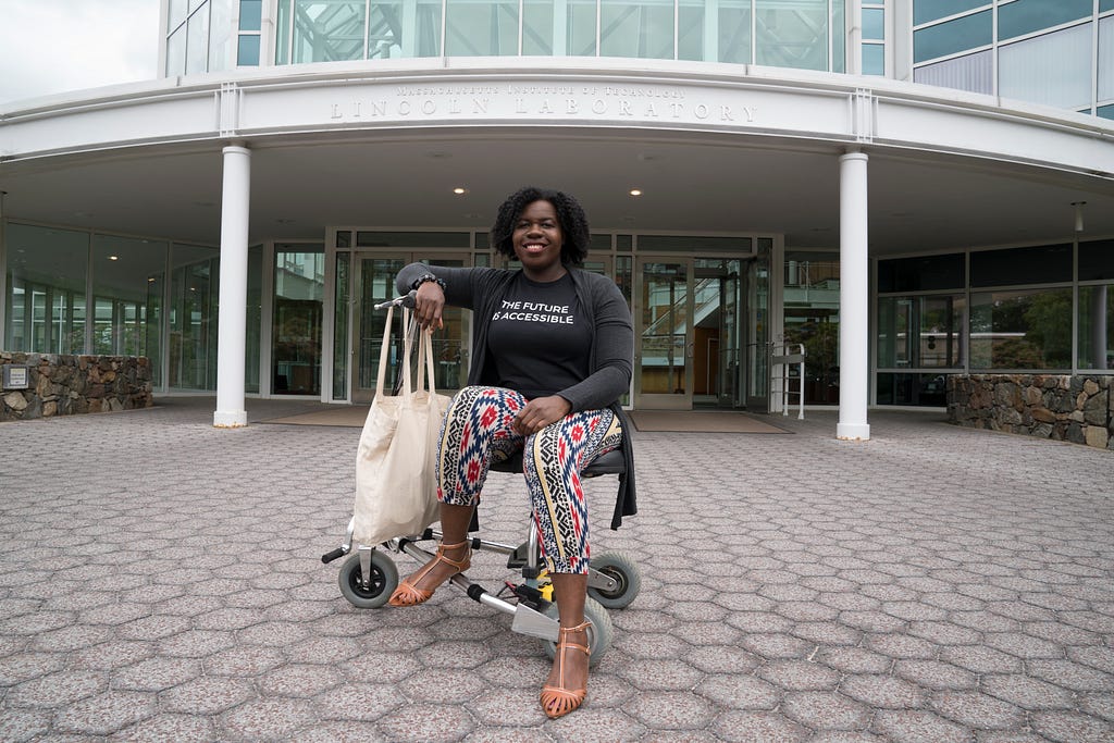 a dark-skinned Black woman seated on a mobility scooter with a black t-shirt that says The Future is Accessible under a grey cardigan, with multicolored trousers
