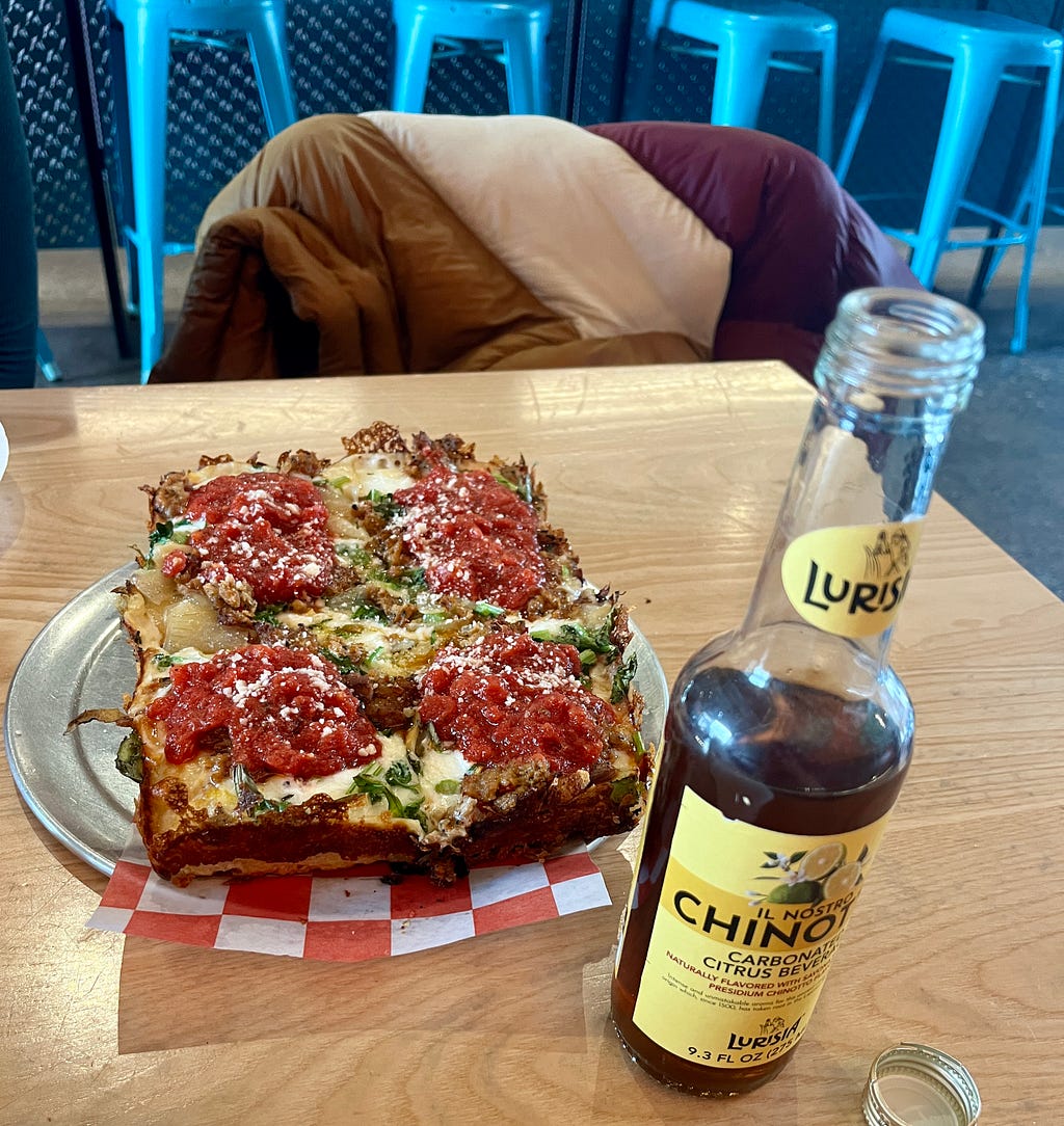 square-shaped pizza and soda bottle on top of a rectangular wooden table, with a brown, cream, and purple jacket hung over a chair behind the table and a row of bright blue bar stools behind the chair.